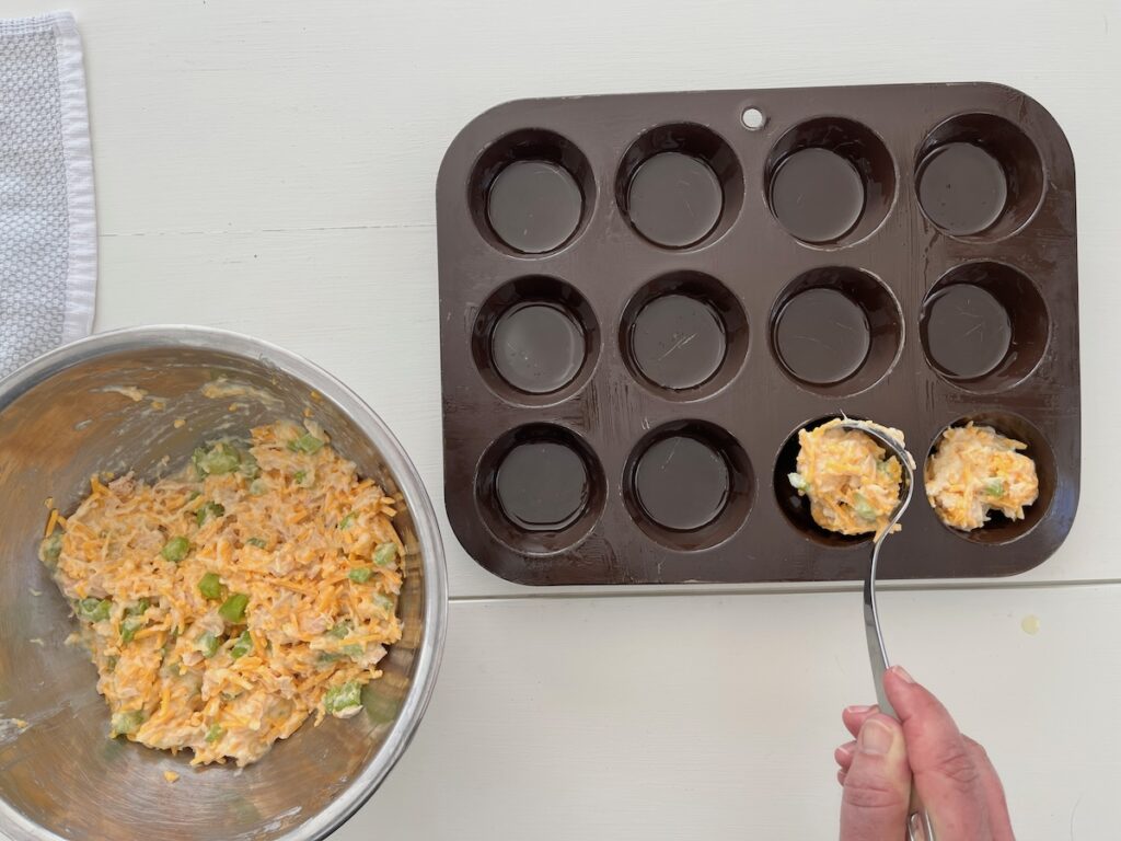 A person fills a muffin tray with a spoonful of a keto tuna melt mixture, rich with shredded cheese and green vegetables.