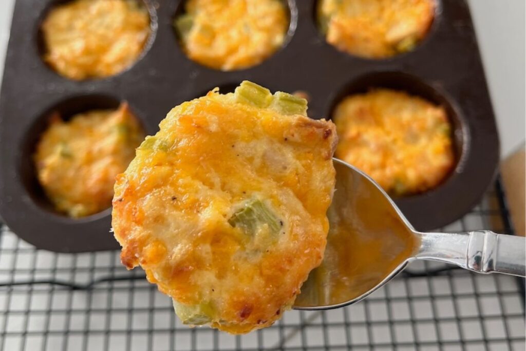 A spoon holds a baked keto tuna melt muffin with melted cheese and green pieces, in front of a tray with similar muffins resting on a cooling rack.