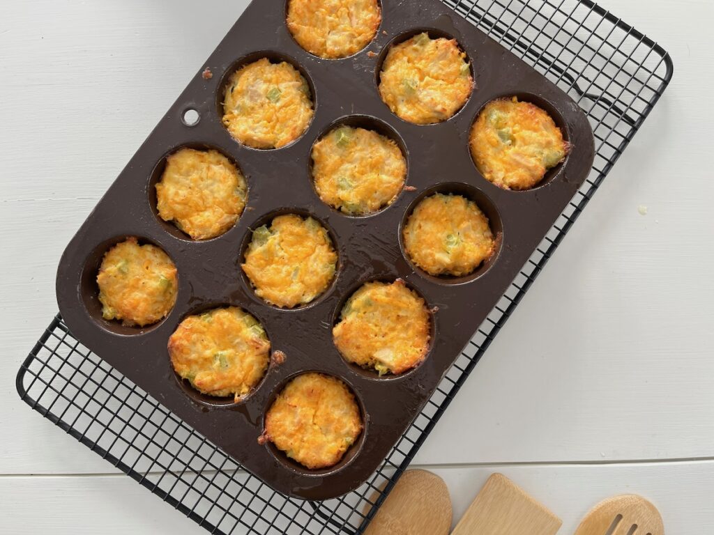 Twelve delicious keto tuna melt-inspired cheese and vegetable muffins sit in a dark muffin tray on a cooling rack, with wooden utensils nearby.