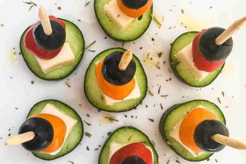 Cucumber slices topped with skewers of black olives, cherry tomatoes, and cheese cubes, garnished with herbs.