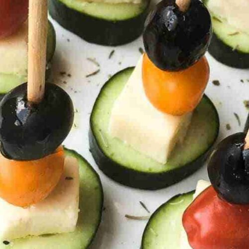 Appetizers with skewers of black olives, cherry tomatoes, and cheese cubes on cucumber slices.