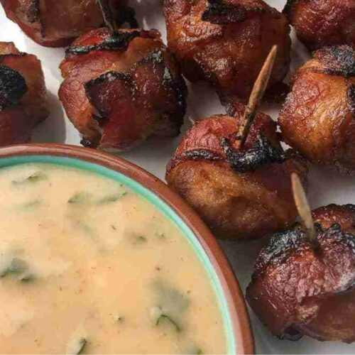 Close-up of bacon-wrapped appetizers on toothpicks beside a bowl of creamy dipping sauce with herbs.