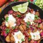 A skillet with baked eggs, red potatoes, crispy bacon, and cilantro, garnished with lime slices. A small bowl of diced avocado is on the side.