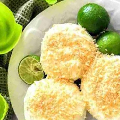 Three round pastries topped with grated coconut on a white plate, garnished with limes, next to green plastic bowls.