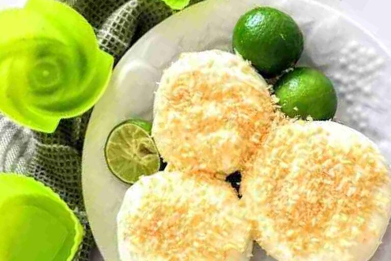 Three round pastries topped with grated coconut on a white plate, garnished with limes, next to green plastic bowls.