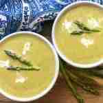 Two bowls of creamy instant pot asparagus soup garnished with cream and asparagus tips rest on a wooden board. A blue patterned cloth adds a touch of charm in the background.