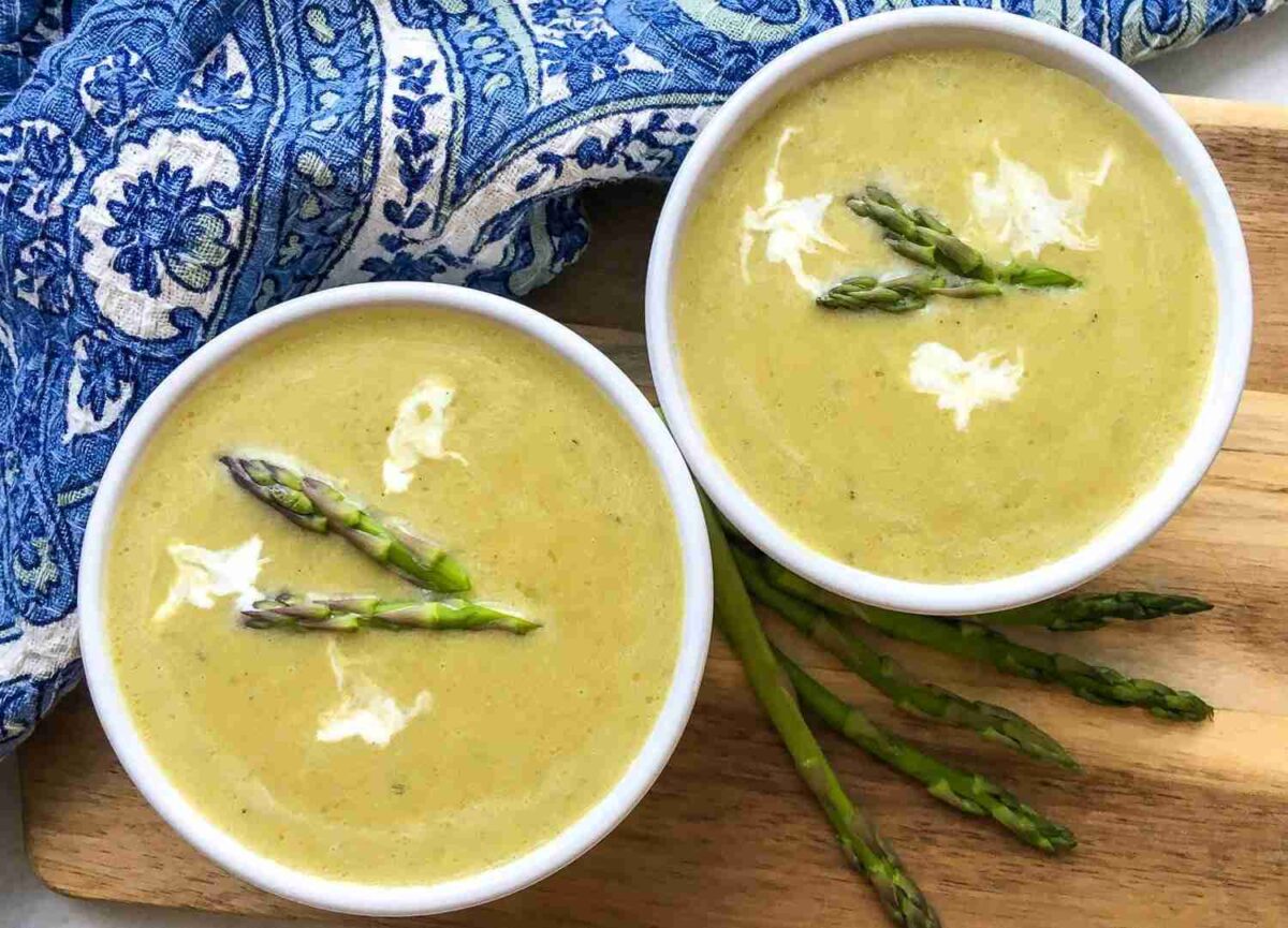 Two bowls of creamy instant pot asparagus soup garnished with cream and asparagus tips rest on a wooden board. A blue patterned cloth adds a touch of charm in the background.