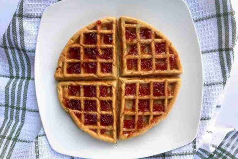 Waffle topped with red jam, divided into four sections, placed on a white plate with a green and white checkered cloth underneath.