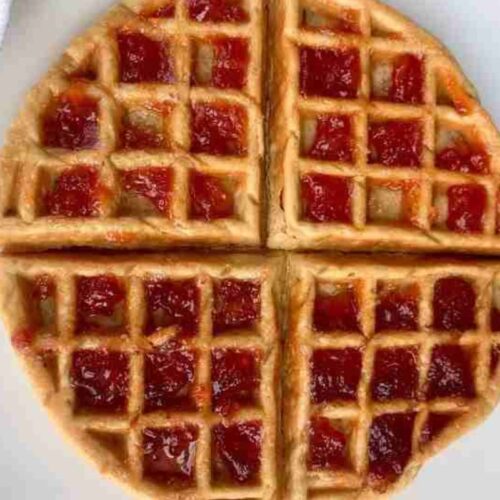 Waffle cut into four quarters, topped with strawberry jam, on a white plate.