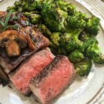 Sliced steak topped with sautéed mushrooms and onions, served with a side of roasted broccoli on a white plate.