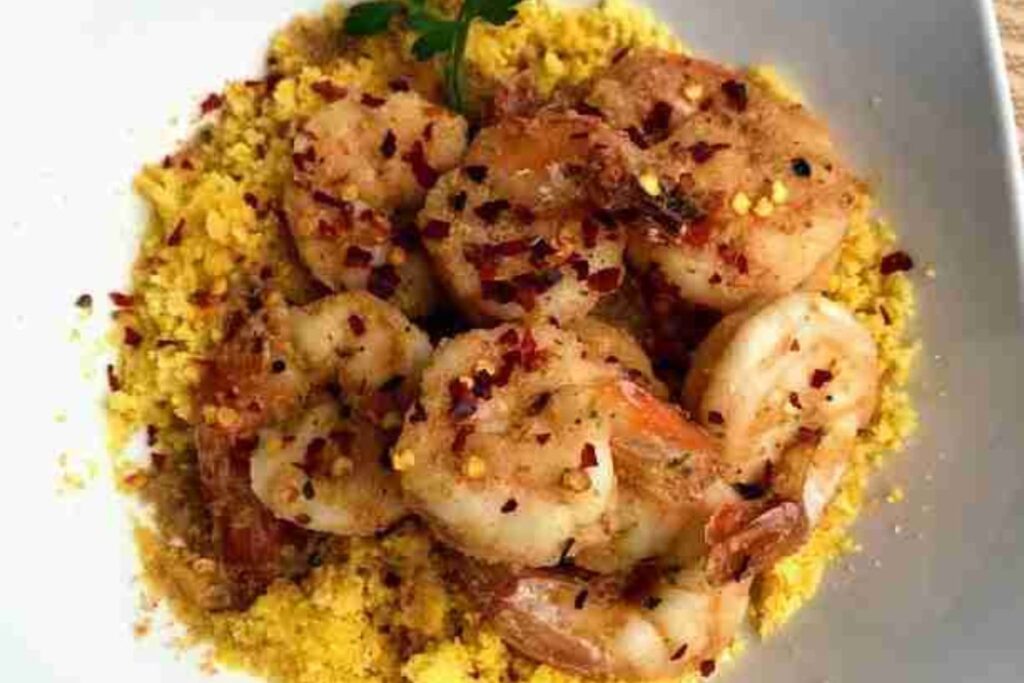 A plate of seasoned shrimp on a bed of golden couscous, garnished with red pepper flakes and a sprig of parsley.
