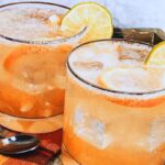 Two glasses of iced orange-colored drink garnished with lemon slices, placed on a tray with a plaid cloth and a spoon beside them.