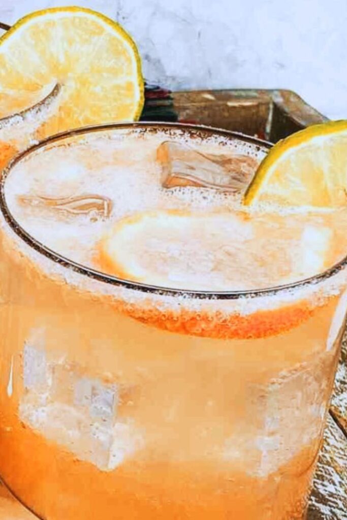 A glass of orange-colored iced beverage garnished with a slice of lime on the rim, sitting on a textured surface.