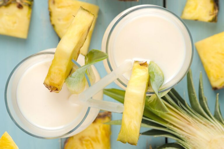 Two glasses of creamy, sugar-free pina colada drinks topped with pineapple slices and straws, surrounded by pineapple pieces on a light blue surface.