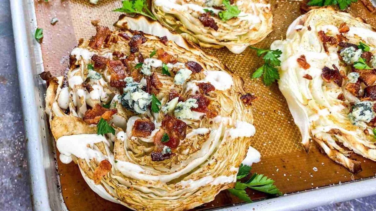 Roasted cabbage wedges topped with chopped bacon, crumbled blue cheese, and fresh herbs on a baking tray.