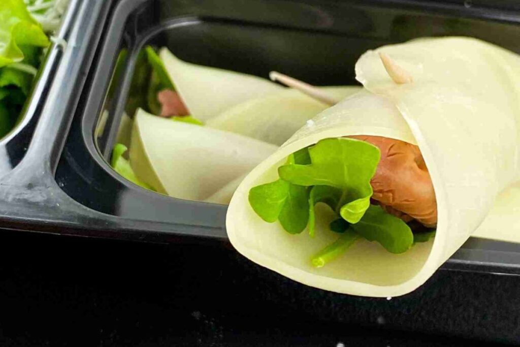 A food container holds a rolled slice of cheese filled with leafy greens and deli meat.