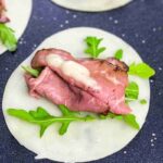 A slice of roast beef with arugula and creamy sauce sits on a circular rice paper wrapper, next to three toothpicks on a dark surface.