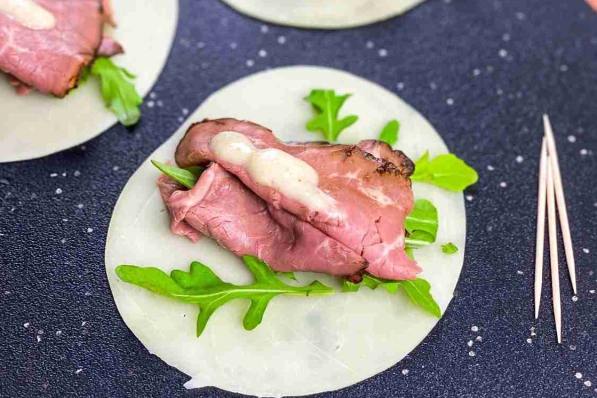 A slice of roast beef with arugula and creamy sauce sits on a circular rice paper wrapper, next to three toothpicks on a dark surface.