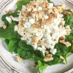 A salad with fresh spinach leaves topped with a mixture of crumbled cheese, walnuts, and what appears to be a creamy chicken salad, served on a beige plate.