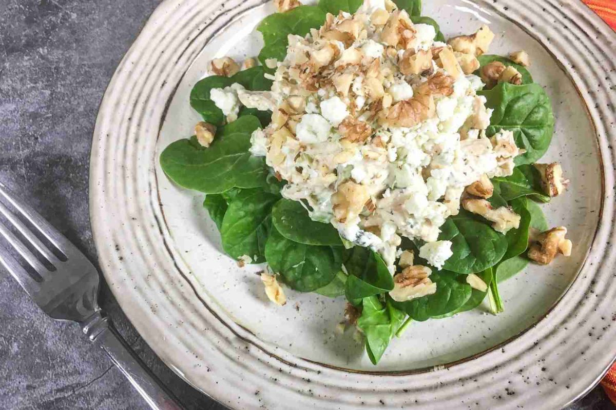 A salad with fresh spinach leaves topped with a mixture of crumbled cheese, walnuts, and what appears to be a creamy chicken salad, served on a beige plate.