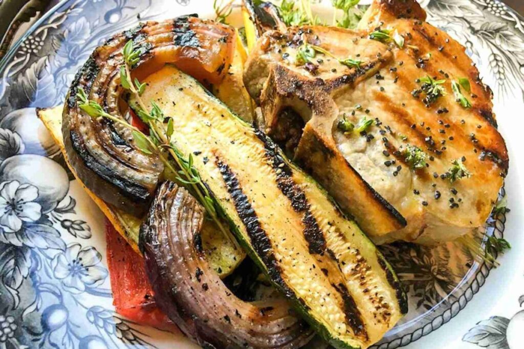 Grilled pork chop with herbs served on a plate with grilled zucchini, onion, and a slice of bell pepper.