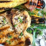 Grilled pork chop garnished with herbs, served with grilled vegetables on a decorative plate.