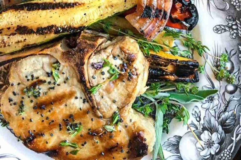 Grilled pork chop garnished with herbs, served with grilled vegetables on a decorative plate.