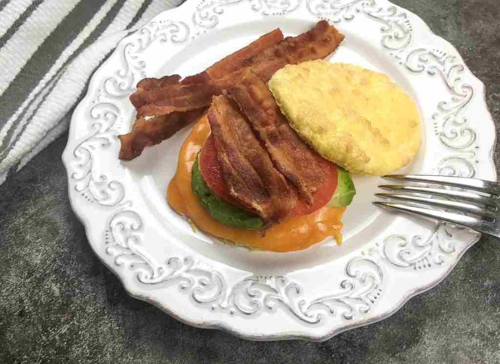 A plate with bacon strips, a cheese, avocado, and tomato sandwich using an egg as the bun. A fork is beside the food.