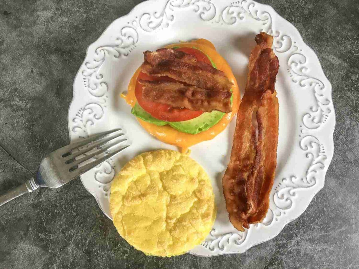 A white plate with bacon, avocado, cheese, tomato, and a round bread, next to a metal fork on a gray surface.