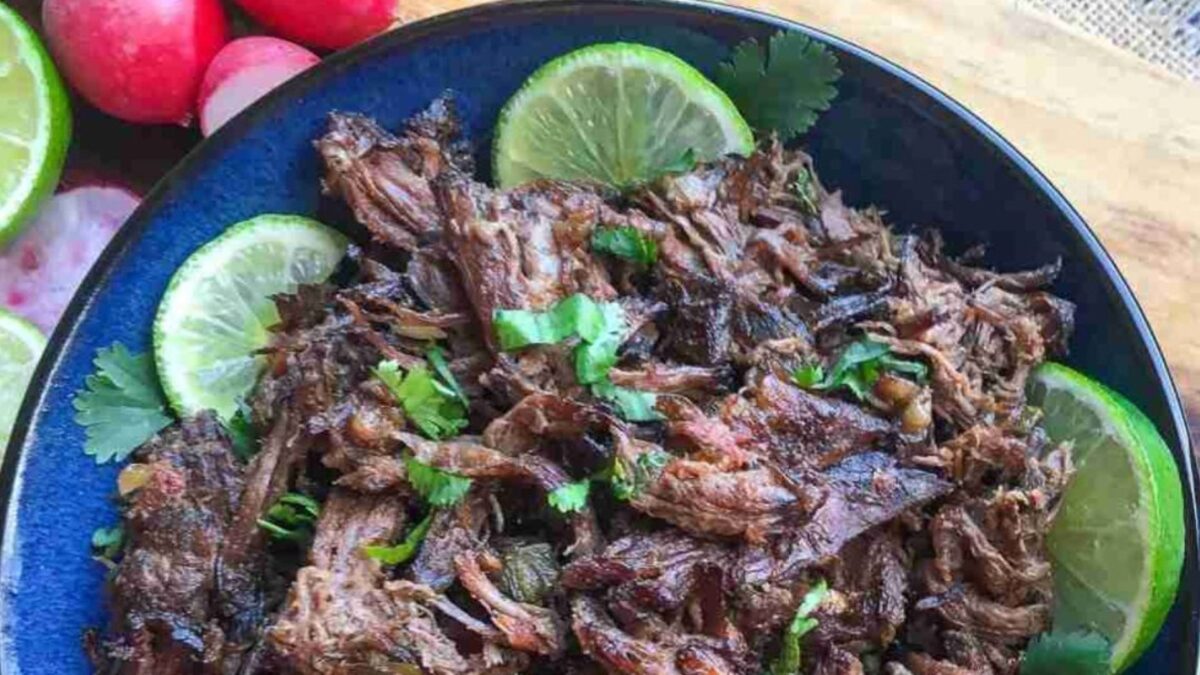 A bowl of slow cooker shredded beef garnished with cilantro and lime slices sits on a wooden surface, accompanied by fresh sliced radishes.
