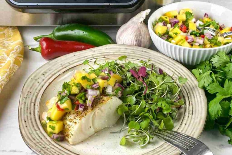 Grilled fish fillet topped with mango salsa on a plate with greens. A bowl of mango salsa, chili peppers, garlic, and cilantro are in the background.