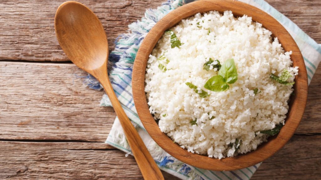Bowl of cauliflower rice garnished with basil leaves on a striped cloth beside a wooden spoon on a rustic wooden surface.