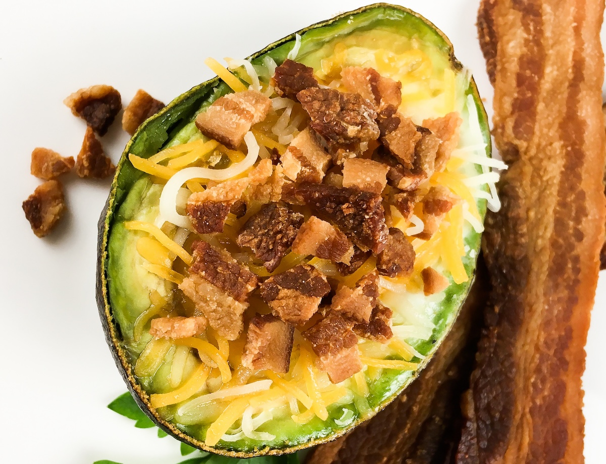 Halved avocado topped with shredded cheese and bacon bits, accompanied by two strips of bacon on the side.