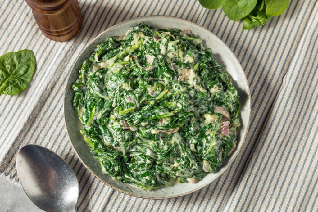 A bowl of creamy spinach salad with visible pieces of diced onion, placed on a striped cloth. A metal spoon and fresh spinach leaves are nearby.