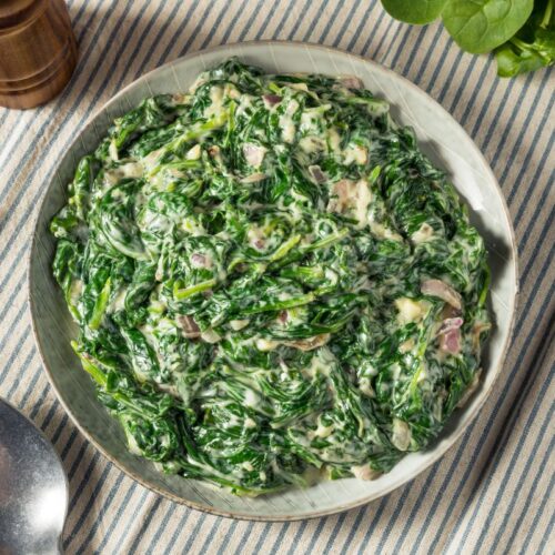 A bowl of creamy spinach salad with visible pieces of diced onion, placed on a striped cloth. A metal spoon and fresh spinach leaves are nearby.