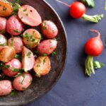 A bowl of roasted radishes garnished with parsley on a dark surface, with three raw radishes and parsley scattered nearby.