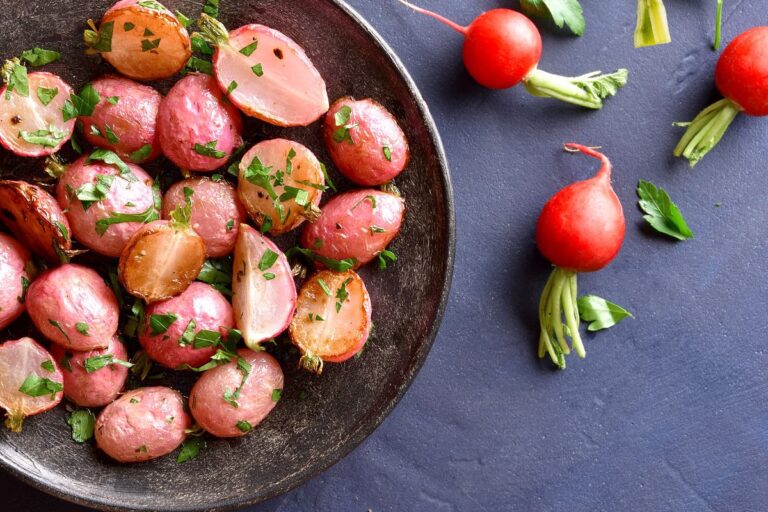 A bowl of roasted radishes garnished with parsley on a dark surface, with three raw radishes and parsley scattered nearby.