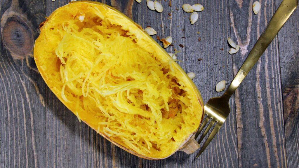 A roasted spaghetti squash half on a wooden surface with a fork and seeds scattered around.