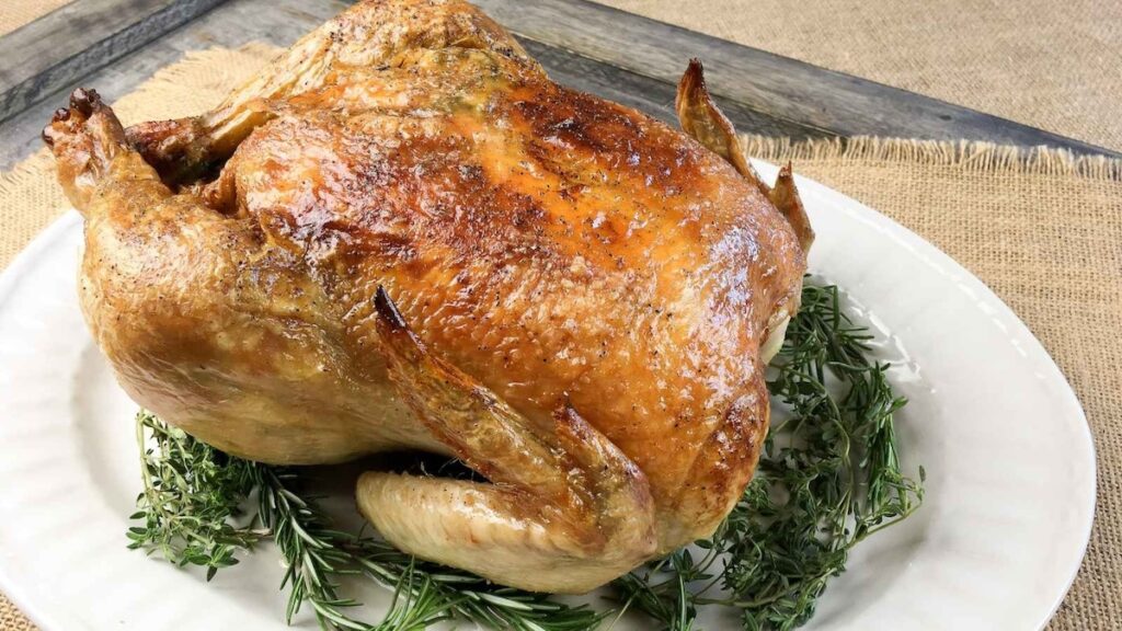 Roasted whole chicken on a white plate, garnished with fresh rosemary, set on a rustic burlap background.
