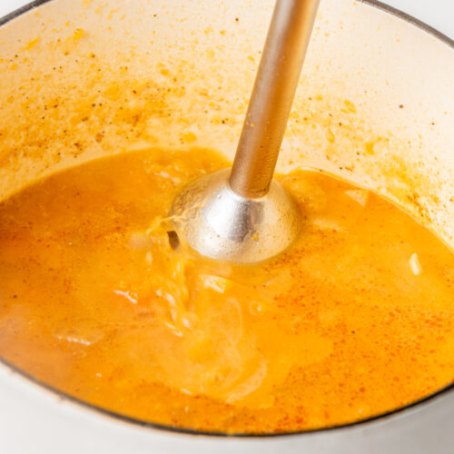 An immersion blender is blending orange soup in a white pot.