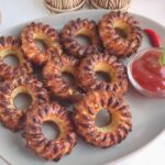 A plate of round, spiced snacks arranged in a circle with a small bowl of red sauce on the side. Two wooden spoons are placed nearby.