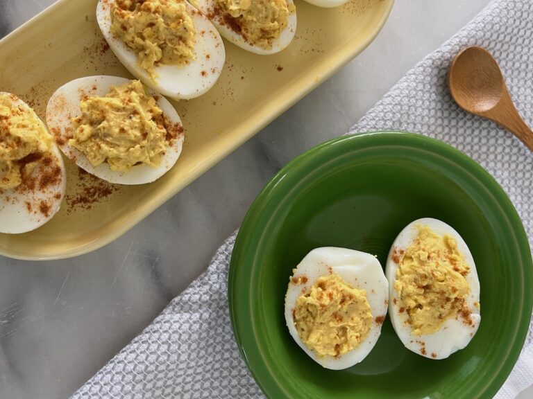 Deviled eggs topped with paprika are served on a yellow platter and a green plate. A wooden spoon is nearby on a textured cloth.