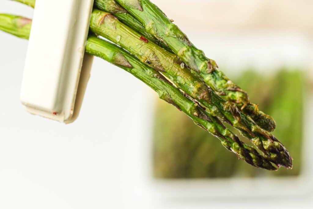 Close-up of fresh green asparagus held by a white clip. The background is blurred with a hint of more asparagus.