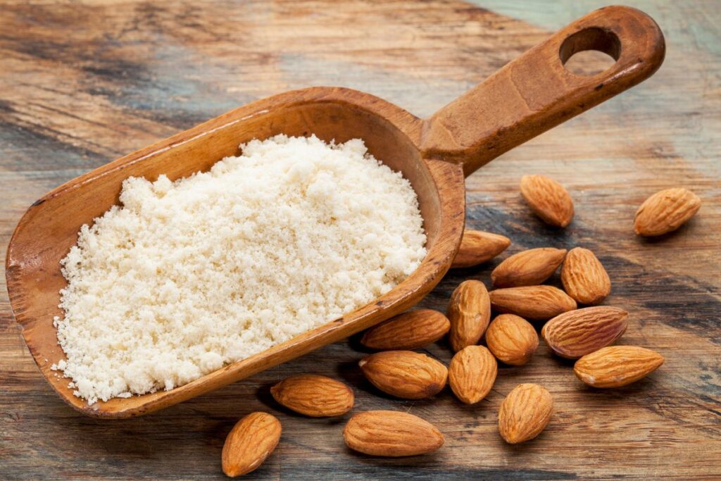 Wood scoop of almond flour surrounded by whole almonds.