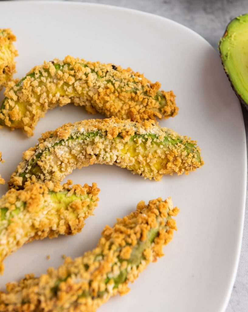 Close-up of crispy avocado fries on a white plate, with a halved avocado in the background.