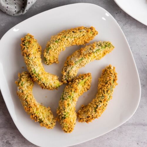 Plate of breaded avocado slices arranged in a circular shape on a white dish, with halved avocados nearby on a gray surface.