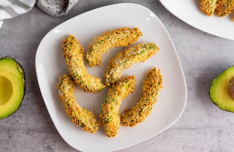 Plate of breaded avocado slices arranged in a circular shape on a white dish, with halved avocados nearby on a gray surface.