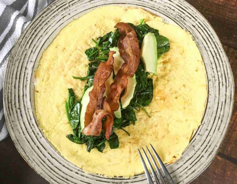 Egg pancake topped with cooked spinach, avocado slices, and two strips of bacon on a plate with a fork on the side.