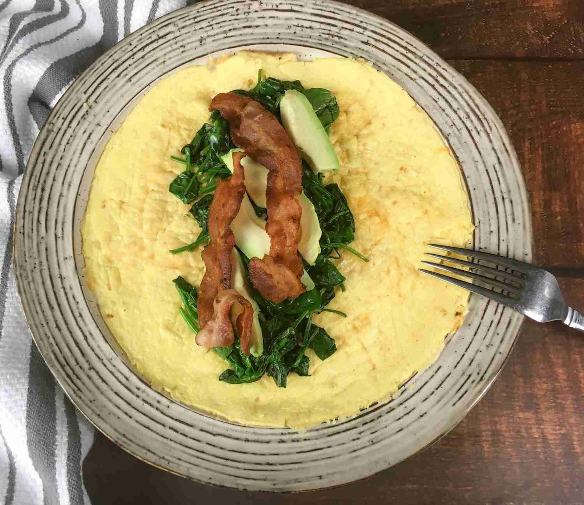 Plate with an open omelette topped with spinach, two strips of bacon, and avocado slices, next to a fork.