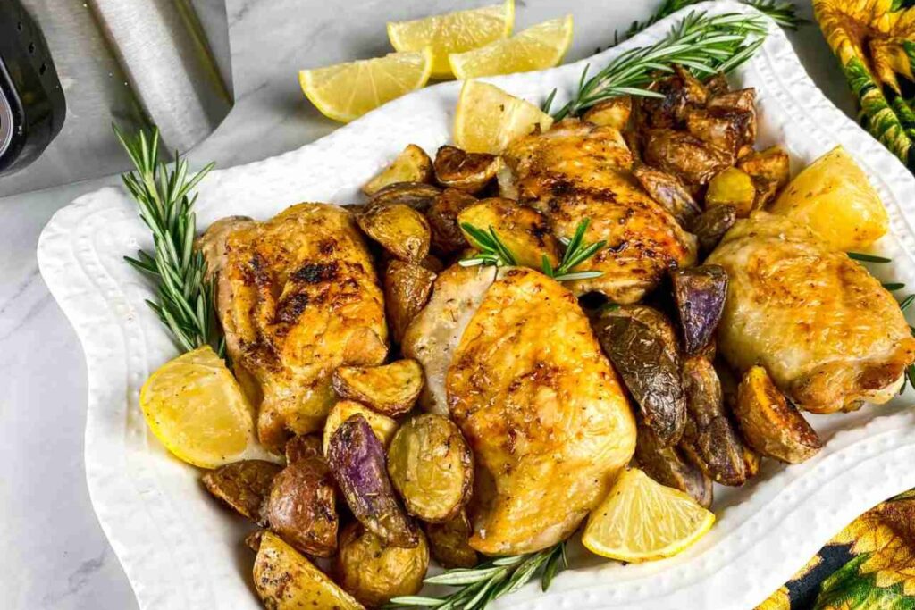 Roasted chicken thighs with assorted potatoes and rosemary garnish on a white platter, accompanied by lemon wedges.